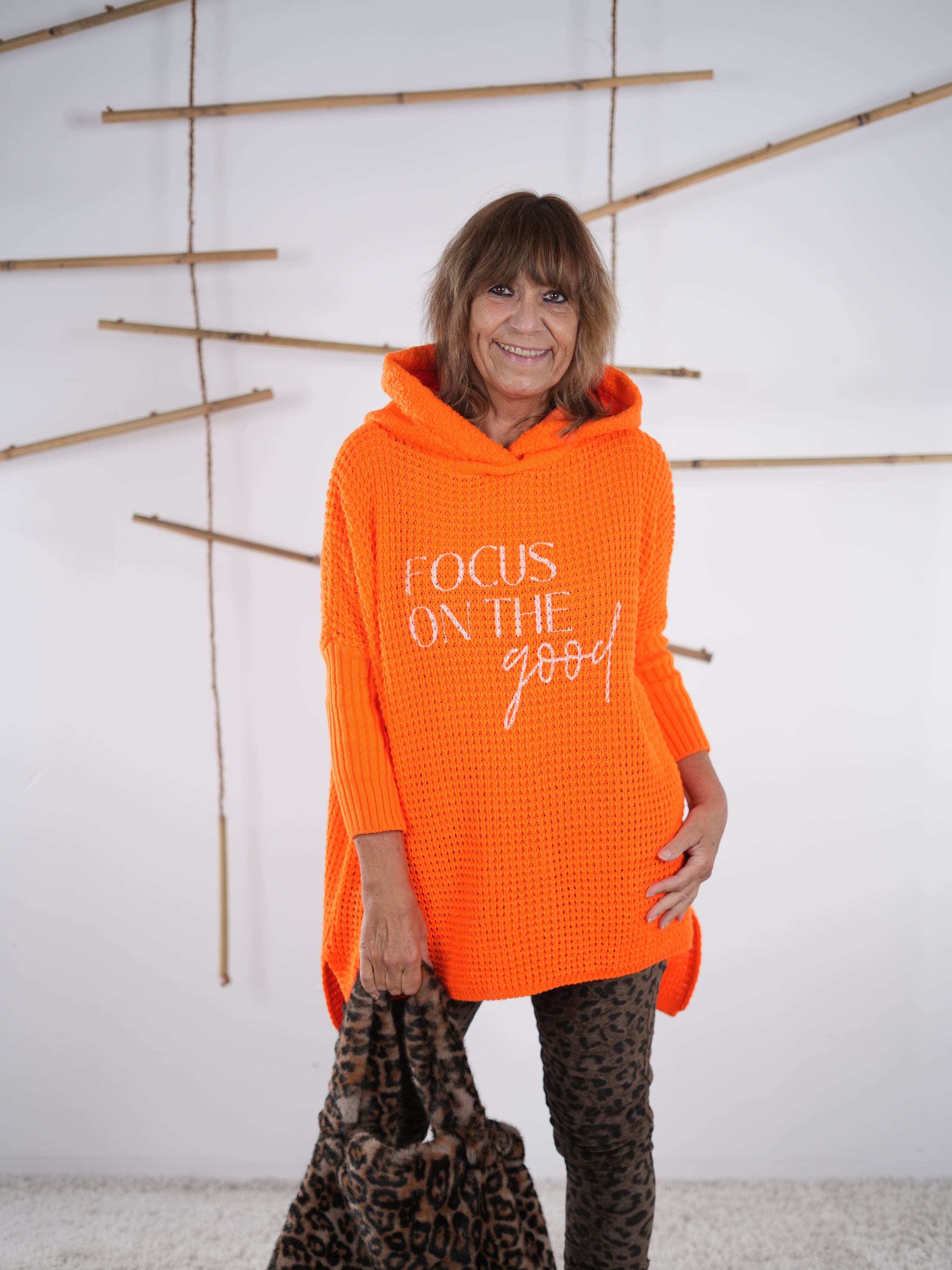 Lässiger Grobstrickpulli in knalliger Farbe mit Schriftzug, Kapuze und Vokuhila-Schnitt in der Frontansicht. Orangene Variante