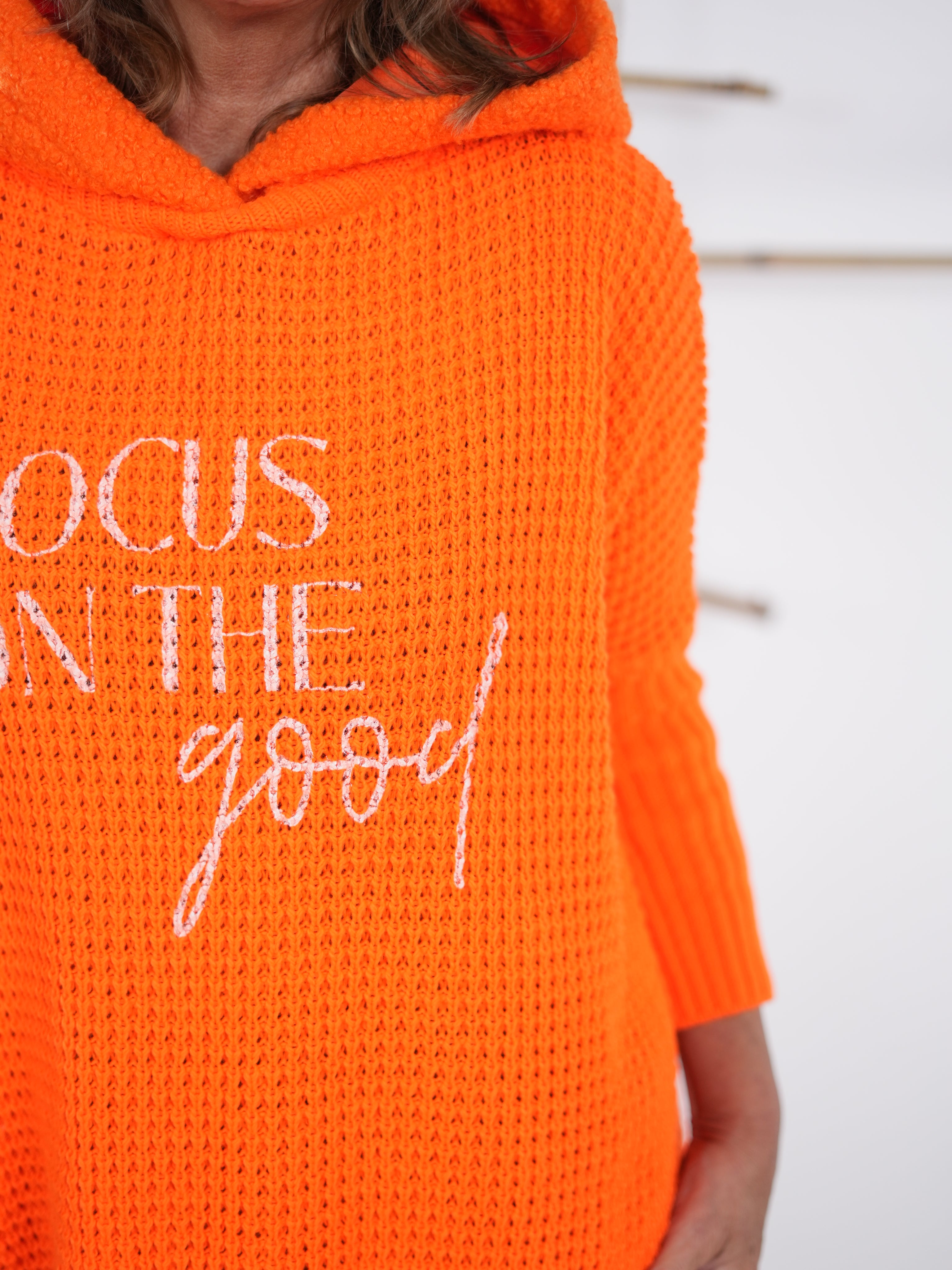 Detailaufnahme des Schriftzugs auf dem orangefarbenen Grobstrickpulli mit grober Maschenstruktur.