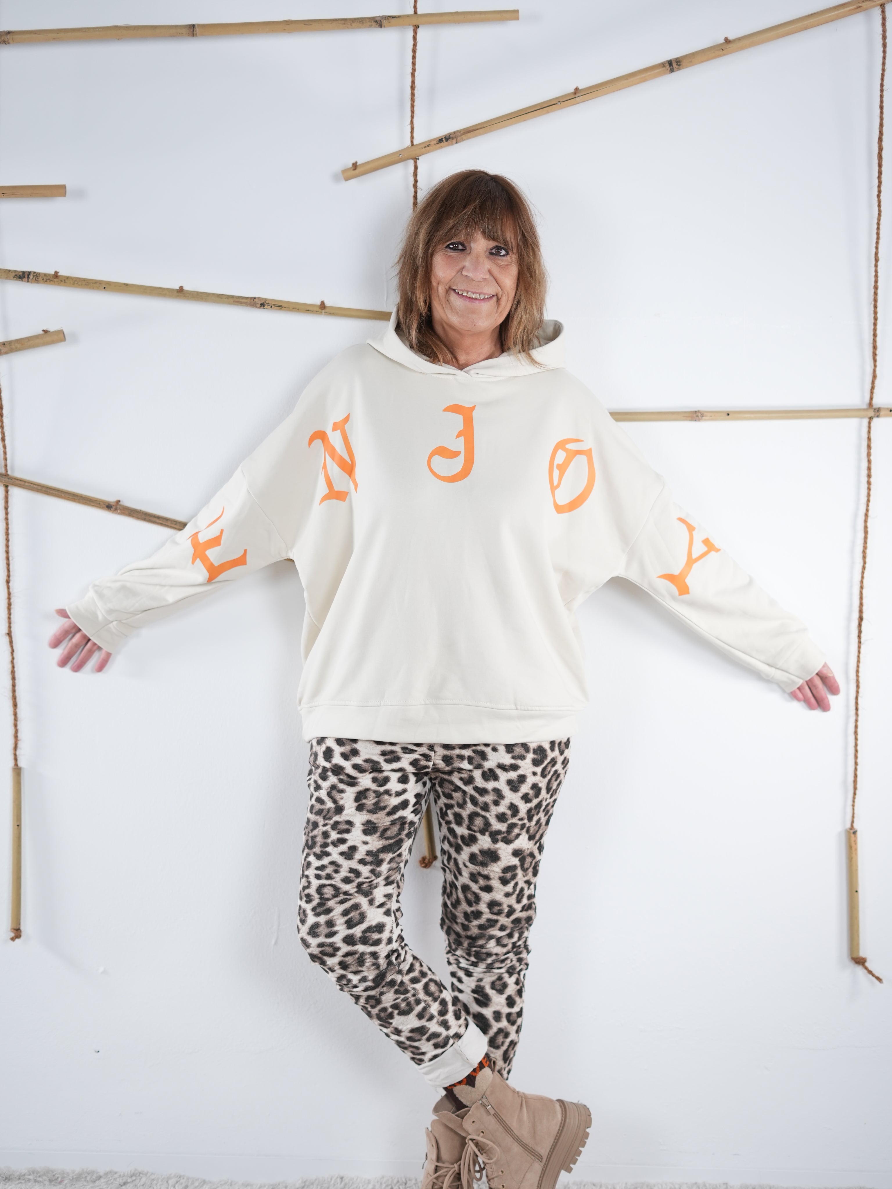 Wollweißer Damen-Hoodie mit großem orangefarbenem ’ENJOY’-Print über Brust und Ärmeln, kombiniert mit einer Leopardhose.