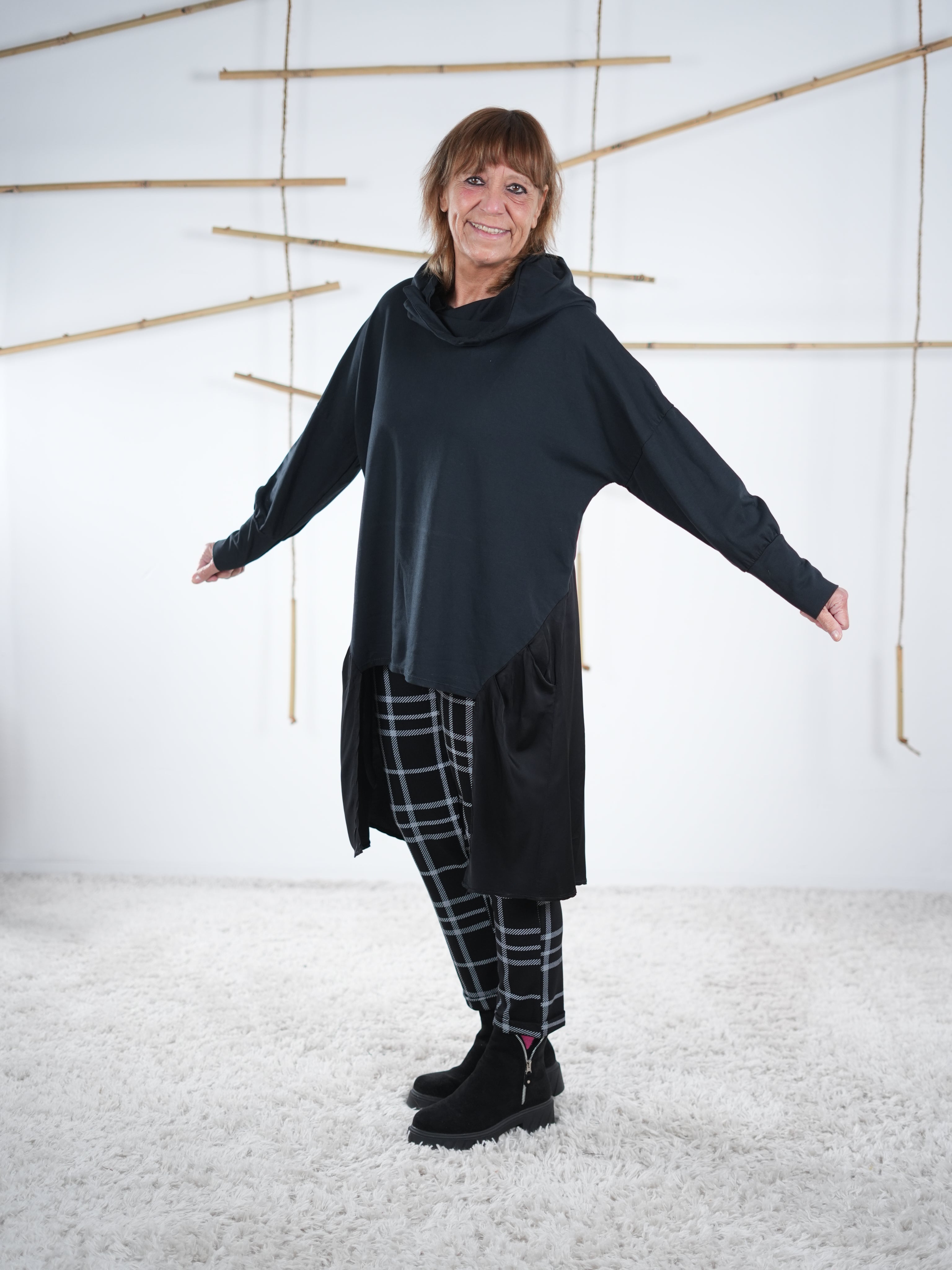 Schwarze Oversize-Tunika mit Satin-Einsatz und Kapuze, Tragefoto mit ausgestreckten Armen.