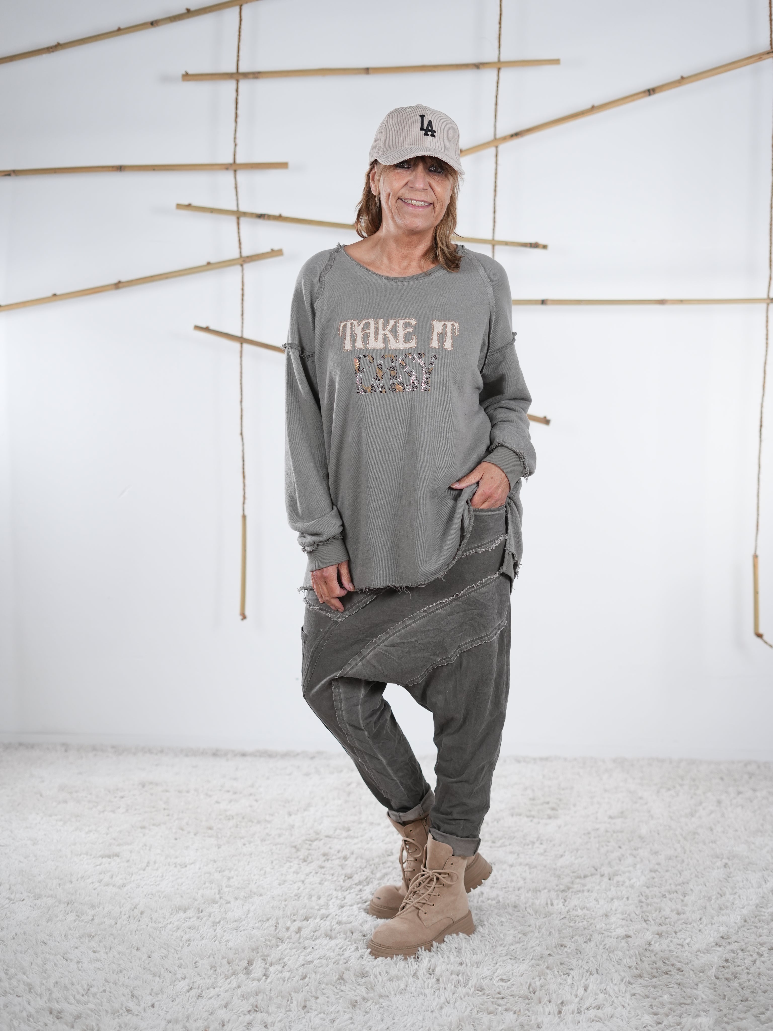 Frau trägt ein taupefarbenes Vokuhila-Sweatshirt mit ‚Take It Easy‘ Schriftzug und grauer Patchwork-Hose.