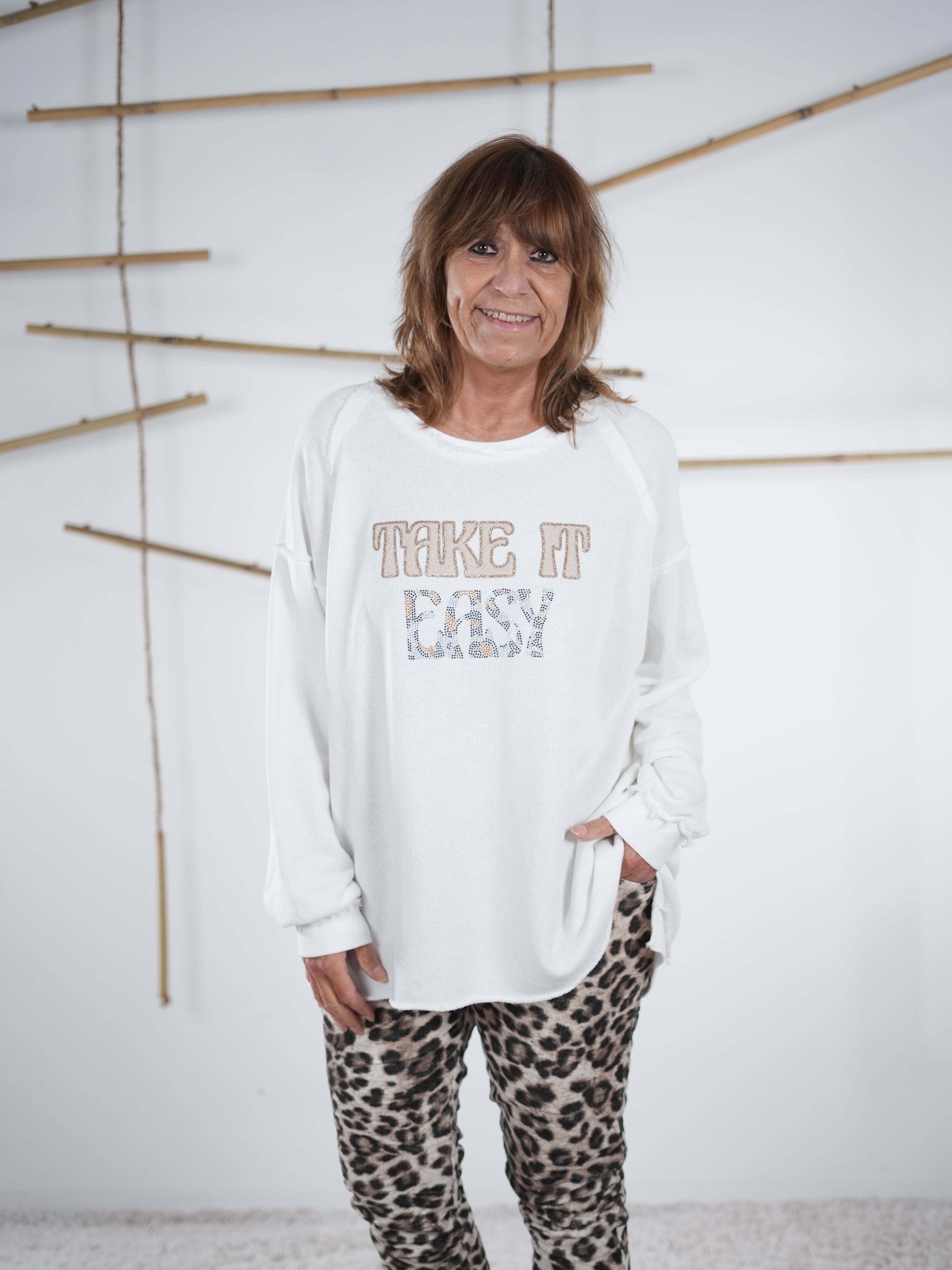Frau trägt ein weißes Vokuhila-Sweatshirt mit ‚Take It Easy‘ Schriftzug und einer hellen Leo-Hose