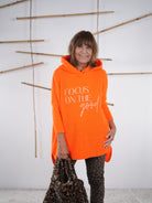 Lässiger Grobstrickpulli in knalliger Farbe mit Schriftzug, Kapuze und Vokuhila-Schnitt in der Frontansicht. Orangene Variante