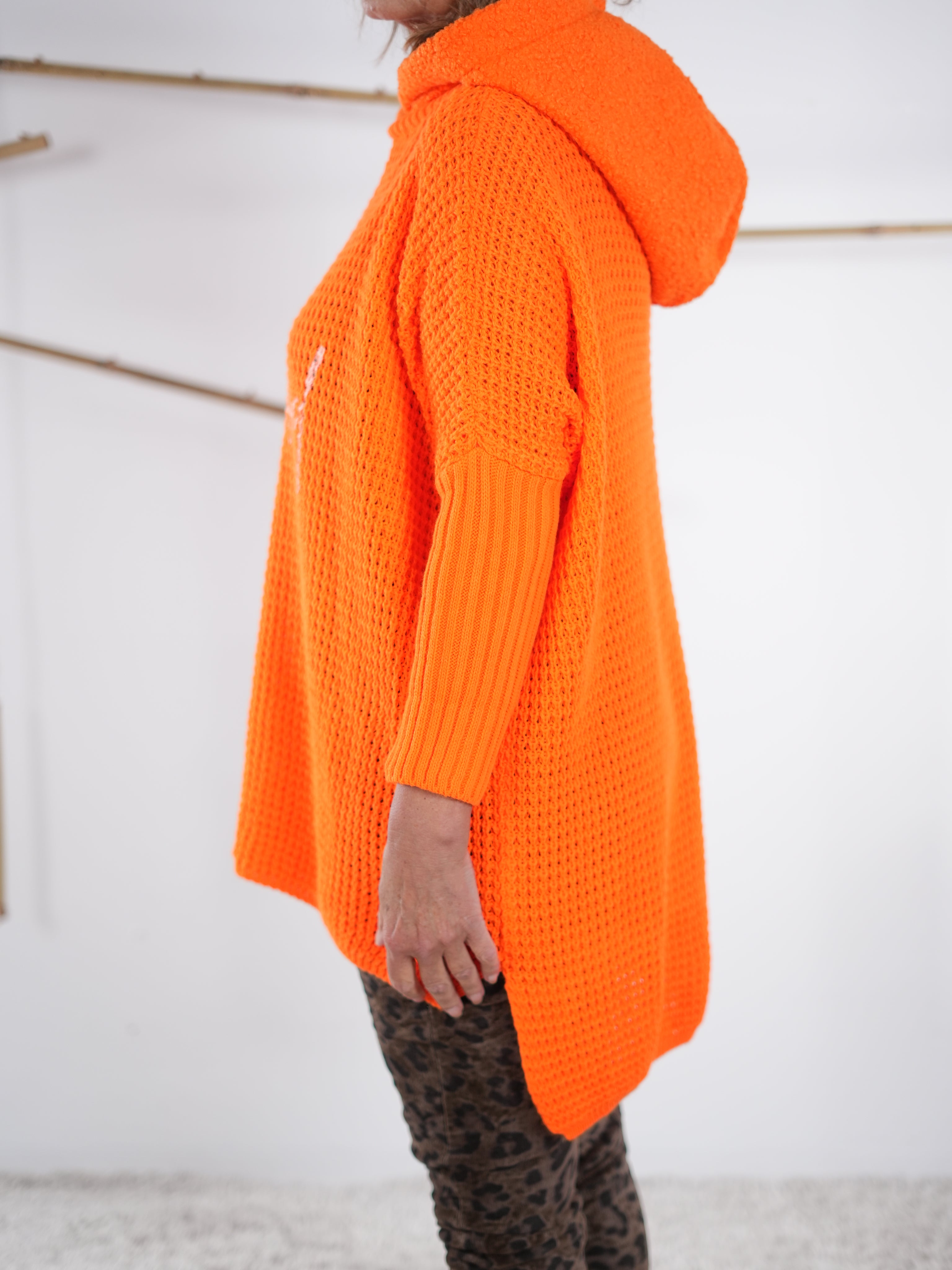 Seitliche Ansicht eines orangefarbenen Grobstrickpullis mit Kapuze und längerem Rückenteil.