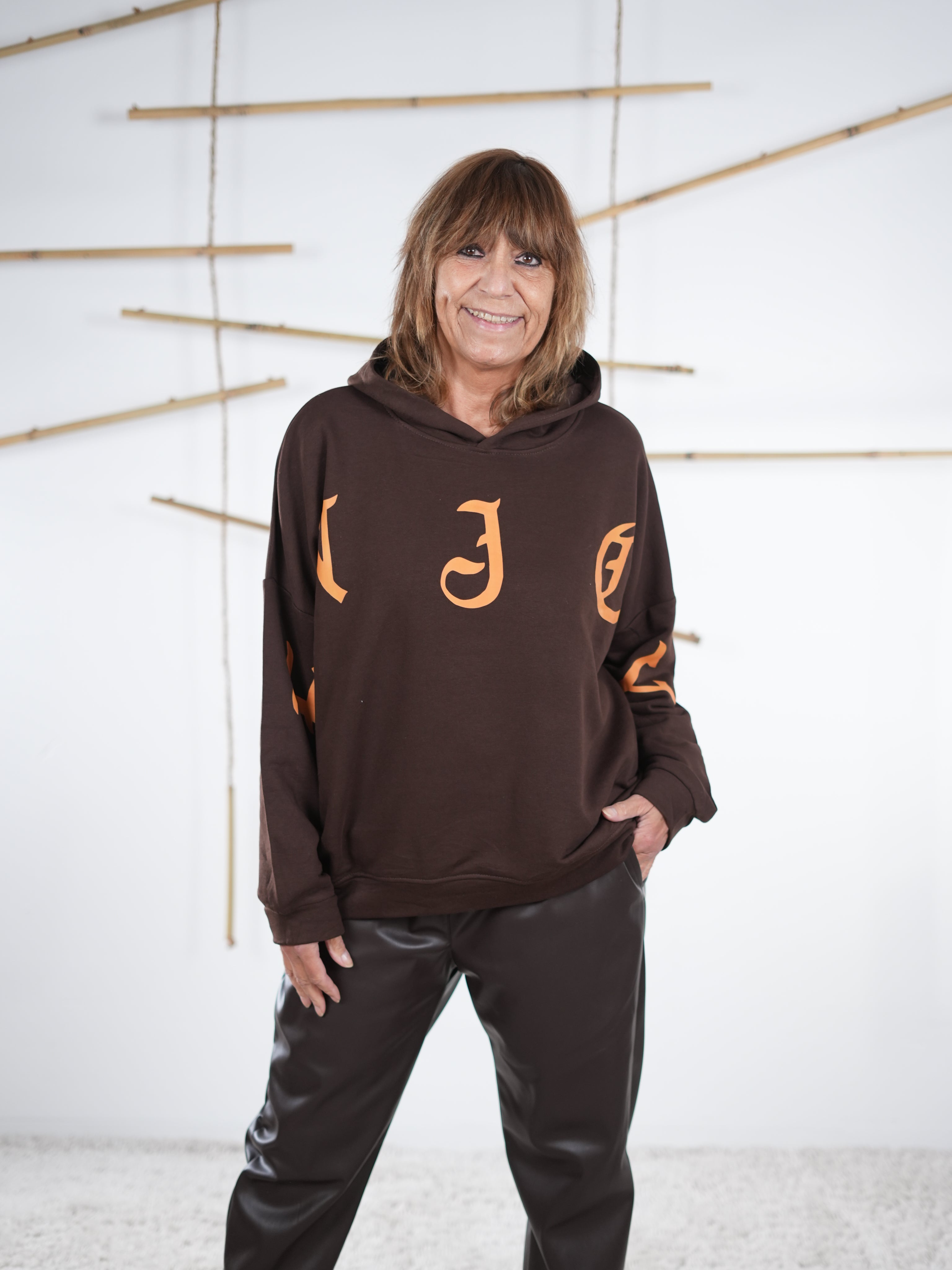Damen-Hoodie in Schokobraun mit großem orangefarbenem Letter-Print vorne, kombiniert mit einer dunkelbraunen Kunstlederhose, mit Kapuze