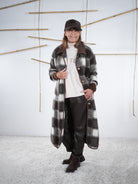 Langer Fleece-Mantel im Karo-Design in Grau-Braun, kombiniert mit hellem Pullover und Lederlook-Hose, frontale Aufnahme.