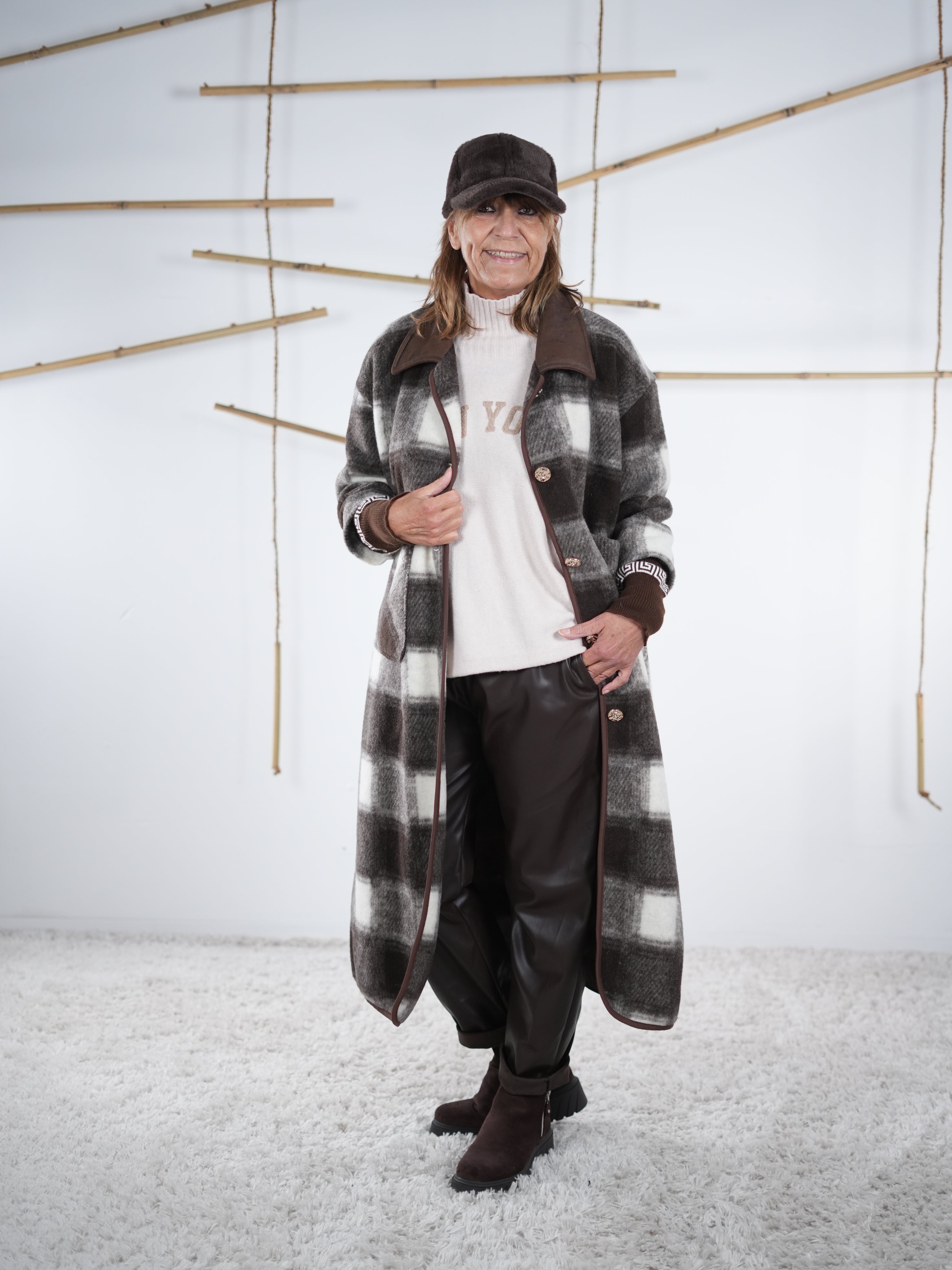 Langer Fleece-Mantel im Karo-Design in Grau-Braun, kombiniert mit hellem Pullover und Lederlook-Hose, frontale Aufnahme.
