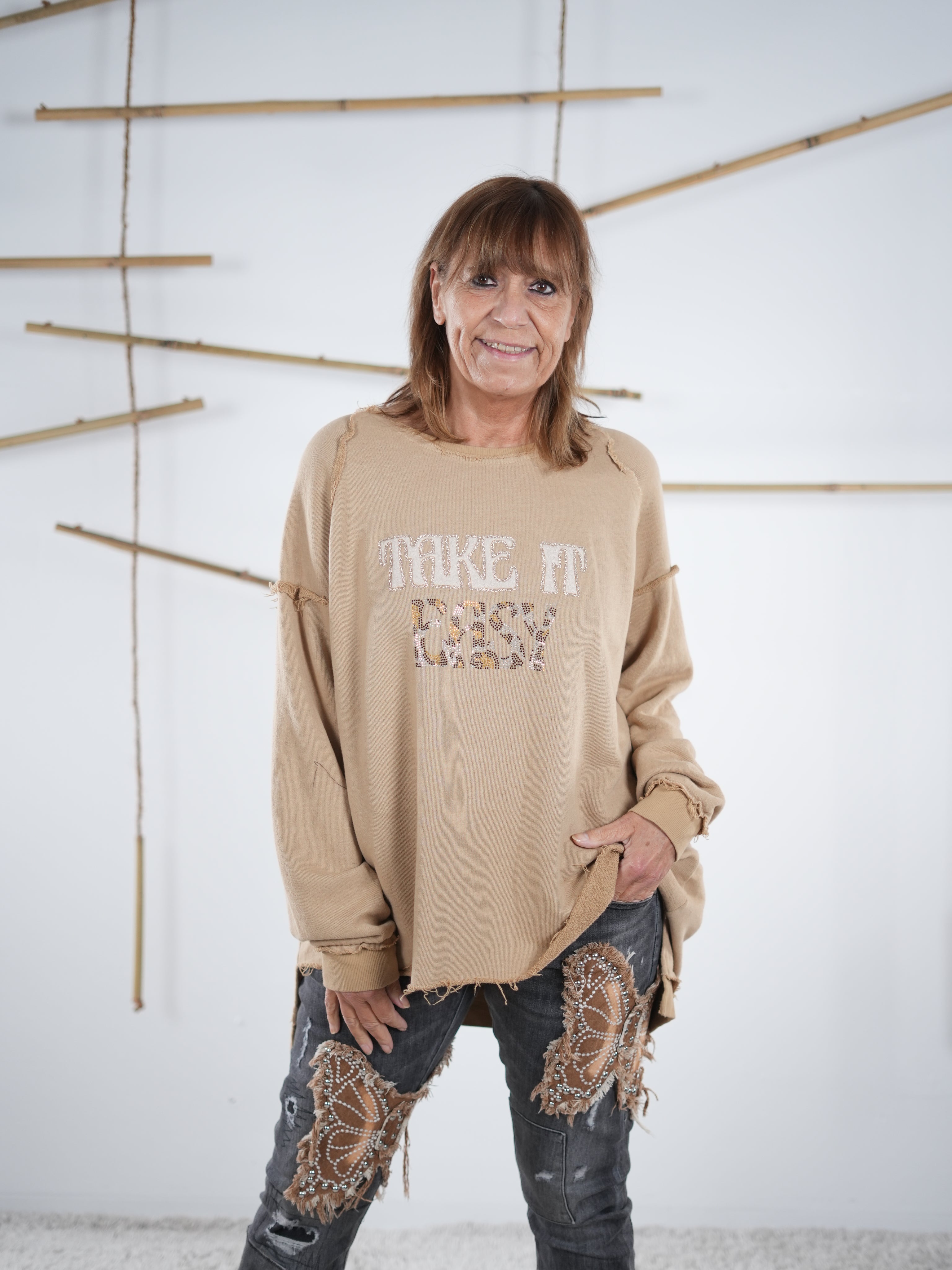 Marion trägt ein camel farbenes Vokuhila-Sweatshirt mit ‚Take It Easy‘ Schriftzug. Der Schriftzug ist mit Leo Akzenten verziert.