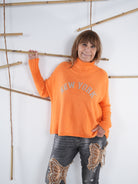 Orangefarbener Damen-Pullover mit „New York“-Schriftzug, Model lehnt seitlich an einer Deko-Bambuswand.
