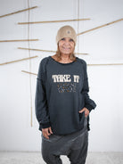 Frau trägt ein anthrazitfarbenes Vokuhila-Sweatshirt mit ‚Take It Easy‘ Schriftzug und legerer grauer Hose.
