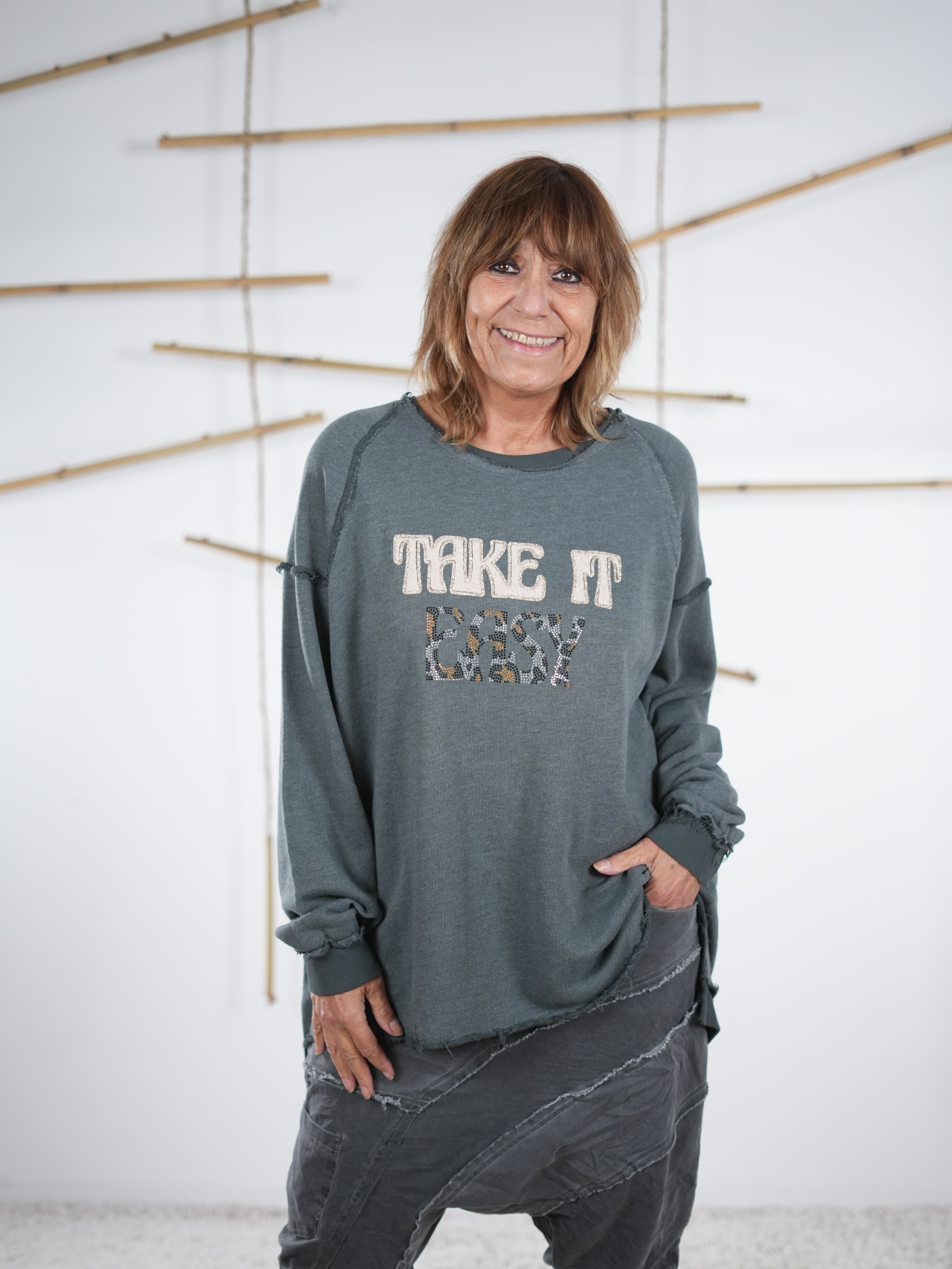 Frau trägt ein graues Vokuhila-Sweatshirt mit ‚Take It Easy‘ Schriftzug und einer grauen Patchwork-Hose.