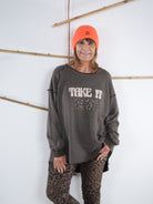 Frau trägt ein schokobraunes Vokuhila-Sweatshirt mit ‚Take It Easy‘ Schriftzug und Leo-Leggings, dazu eine neonorange Mütze.