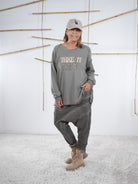 Frau trägt ein taupefarbenes Vokuhila-Sweatshirt mit ‚Take It Easy‘ Schriftzug und grauer Patchwork-Hose.